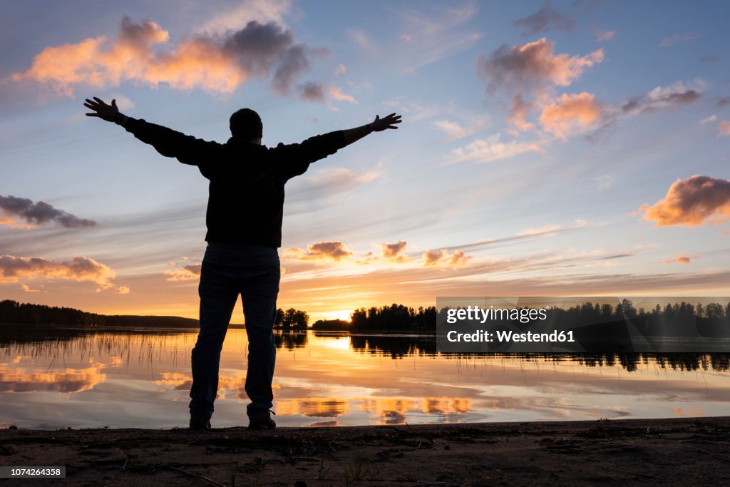 Finland, Kajaani, Man watching sunset at the river with arms outstretched