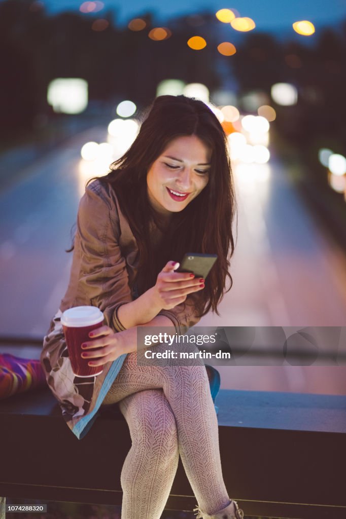 Woman in the city by night drinking coffee and texting