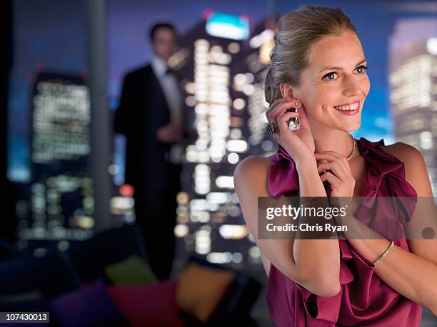 elegant woman putting on earrings with husband in background - women of penthouse stock pictures, royalty-free photos & images