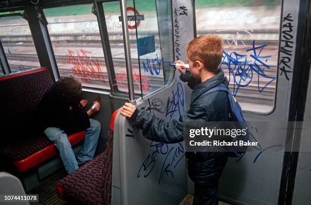 Seen from behind, two young boys tag the inside the 1980s carriage of a 1990s London Underground train, on 8th November 1989, in London, England. In...