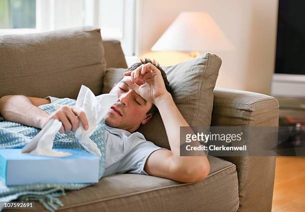 man with a cold lying in sofa holding tissues - cold illness stock pictures, royalty-free photos & images