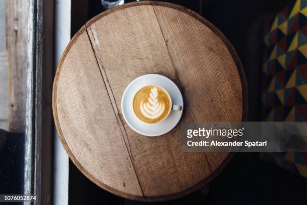 high angle view of coffee cup with latte froth art on round wooden table in cafe - overhead view photos et images de collection