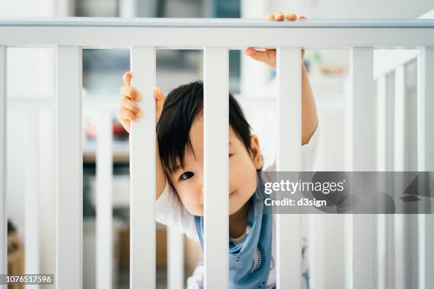 curious baby girl standing in her crib and looking through bars - sicherheitsgitter stock-fotos und bilder