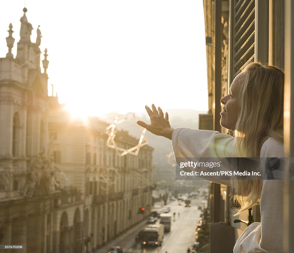 Woman strecthes arms out apartment window, sunrise