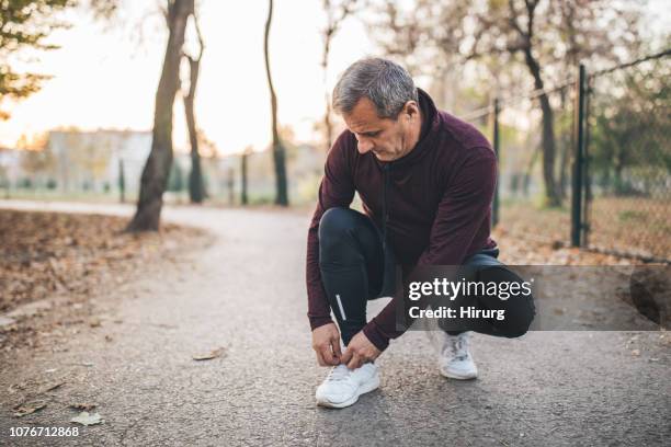 senior man taking pause from jogging to tie his shoelace - mens footwear stock pictures, royalty-free photos & images