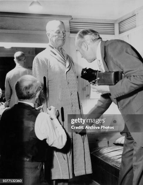 English fashion designer Cecil Beaton fits French actor and singer Maurice Chevalier with a coat on the set of the MGM film 'Gigi', circa 1958.