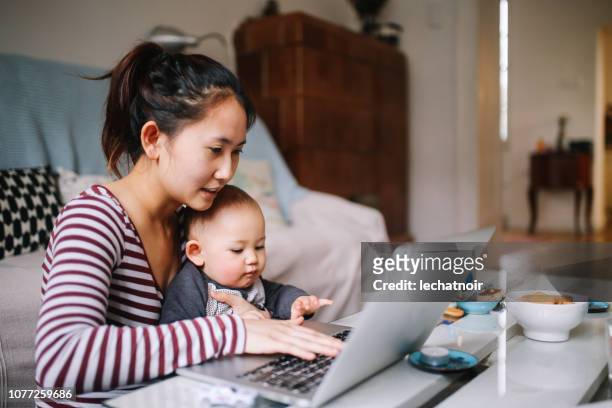giovane mamma asiatica che cerca di lavorare con il suo bambino - familie laptop foto e immagini stock