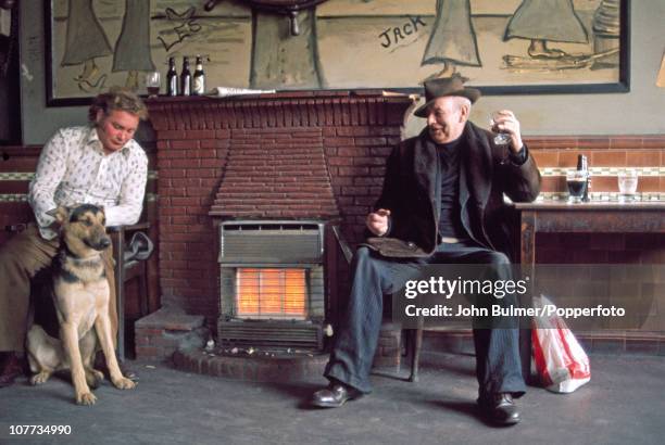 Two men and their dog warm up by a gas fire at a pub in Manchester, England in 1976.