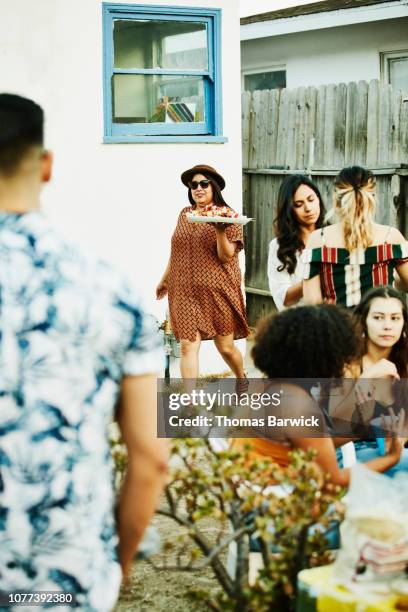 smiling woman bringing plate of food to barbecue during backyard party with friends - day california arrivals stockfoto's en -beelden