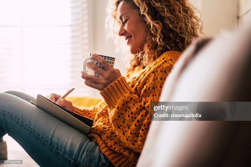 Happy cute lady at home write notes on a diary while drink a cup of tea and rest and relax taking a break. autumn colors and people enjoying home lifestyle writing messages or lists. Blonde curly beautiful lady sit down in the house