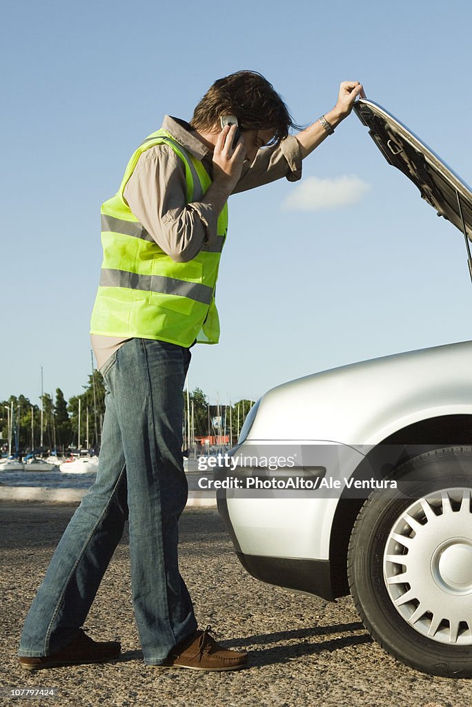 Man making phone call with cell phone while evaluating car broken down on side of road