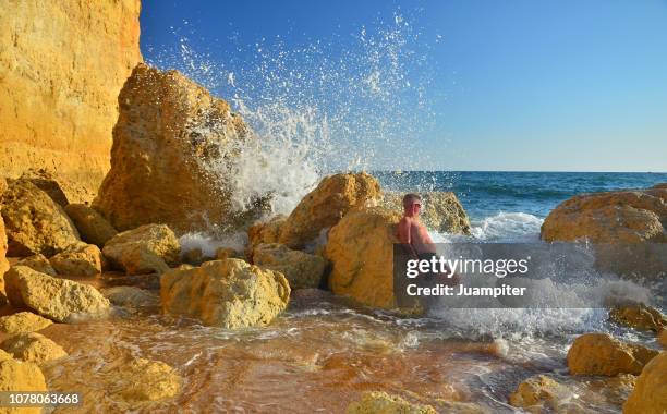 hombre joven disfruta del sol y la playa apoyado en unas rocas mientras una ola rompe sobre él - hombre joven stock pictures, royalty-free photos & images