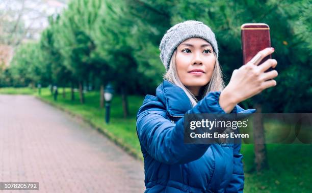 woman taking selfie by mobile phone's camera in public park - blue coat stock pictures, royalty-free photos & images