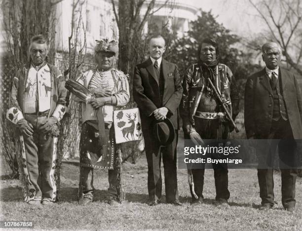 President Calvin Coolidge posed, full-length portrait, standing, facing front, with four Osage Indians, White House in the background, 1925.