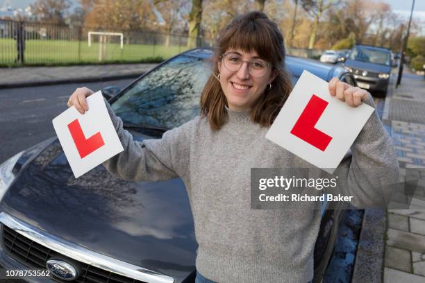 Young 23 year-old woman celebrates the passing of her driving test by holding up her L Plates in front of the family car in south London, on 7th...