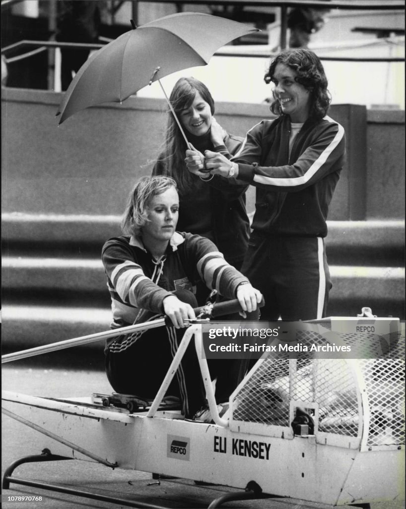 Sally Harding from N.S.W., the stroke of the Australian Four, demonstrates on the ergometer, a machine which is used to gauge strength and output of a rower over a distance. Sally is sheltered from the inclement weather by her team mates Jilliam McCure of
