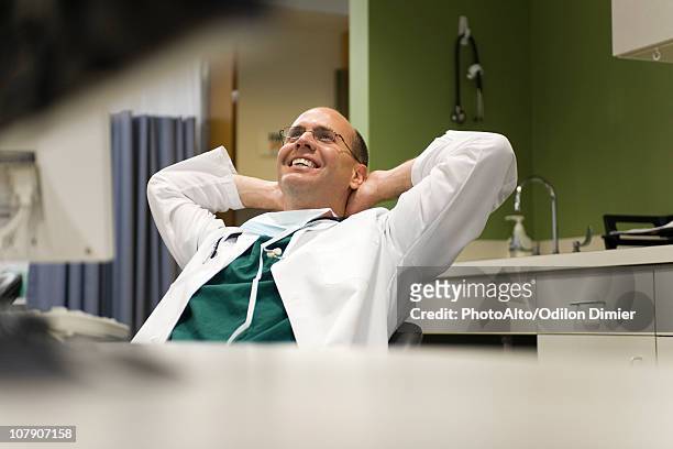 doctor leaning back in chair with hands behind head, smiling - médecin content photos et images de collection
