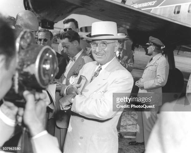 American President Harry S. Truman smiles as he checks his watch and looks into a camera lens while on the tarmac next to his plane, 'Independence,...
