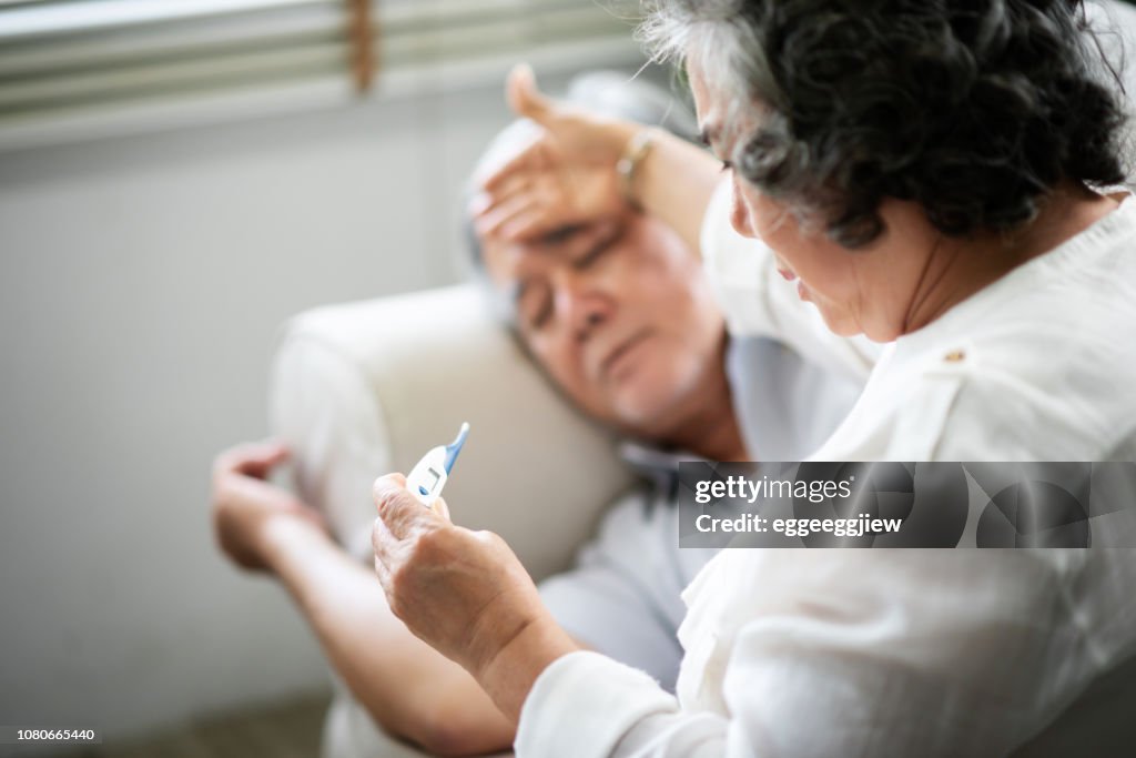 Asiatische Senior Mann auf Sofa, während seine Frau halten und auf der Suche nach Thermometer liegen.