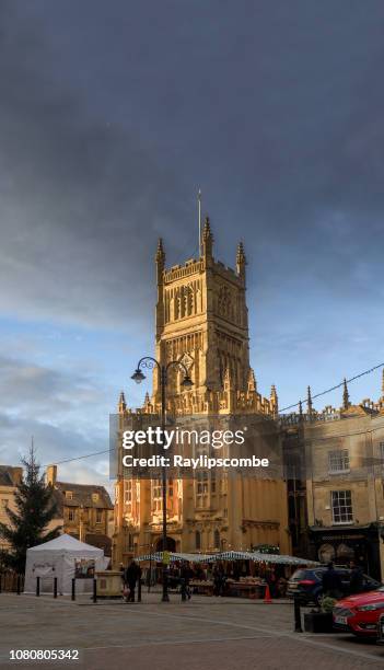 shoppers wandering around market stalls in cirencester, the cotswolds, beneath the parish church of st john the baptist bathed in an evening winter sunset - cirencester stock pictures, royalty-free photos & images