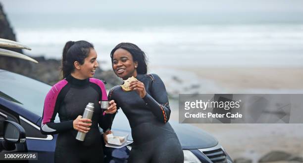 two women having food by car after surfing - beach hold surfboard stock-fotos und bilder