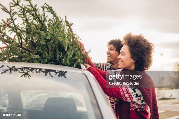 smiling couple tying christmas tree to a car - xmas car stock pictures, royalty-free photos & images