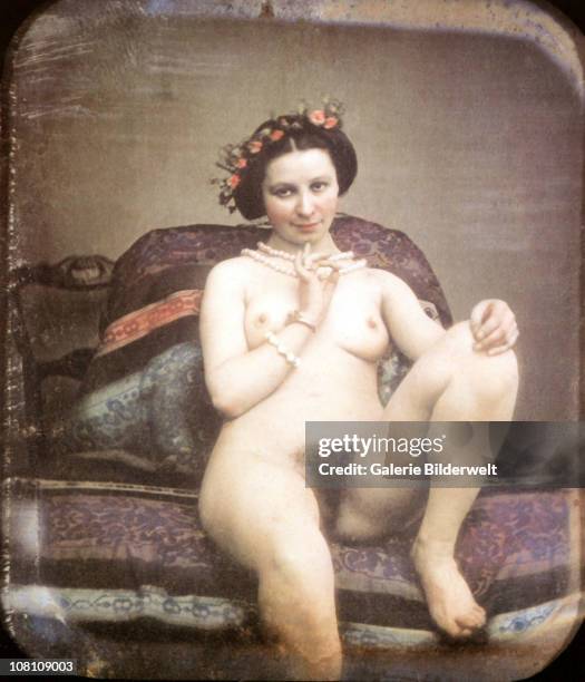 Nude woman is sitting on a chaise-longue wearing nothing but a pearl necklace and armband as well as red flowers in her hair. 1850. Hand-colored...