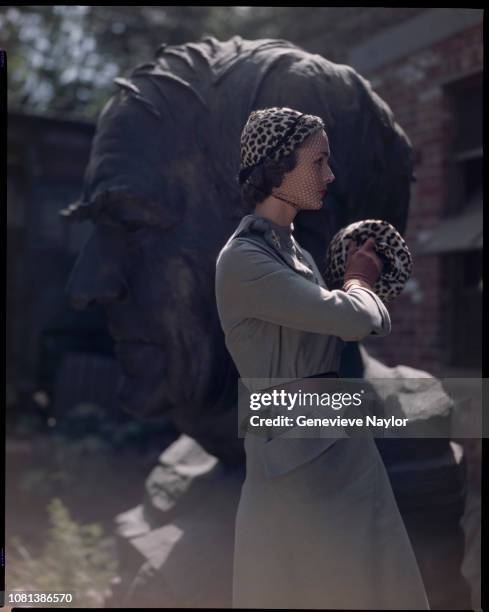 Fashion model wears a leopard hat and muff by Bruno with a suit dress by Hattie Carnegie next to a giant bust of T.A. Edison.