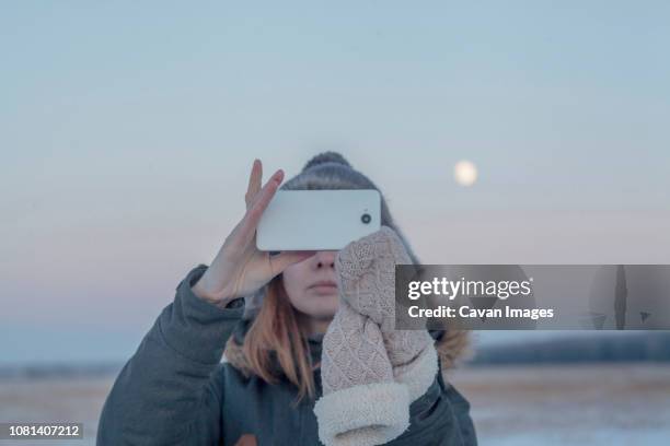 woman photographing with smart phone against sky during sunset - op handschuh stock-fotos und bilder