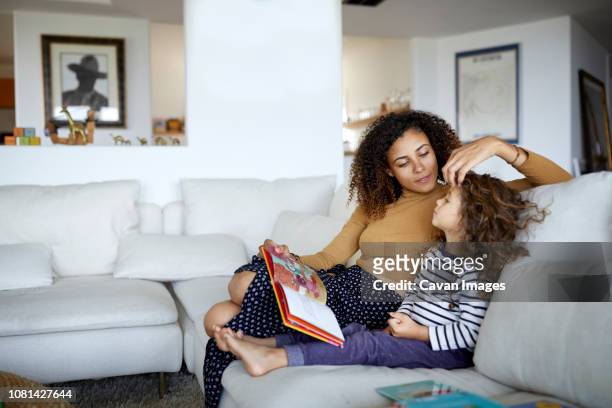 mother reading picture book for cute daughter while sitting on sofa at home - child sitting stock-fotos und bilder