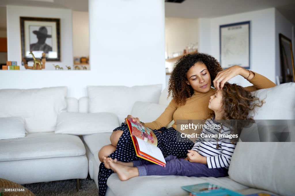 Mother reading picture book for cute daughter while sitting on sofa at home