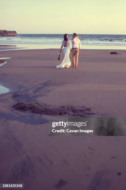 rear view of newlywed couple walking at beach against clear sky during sunset - brautpaar rückenansicht stock-fotos und bilder