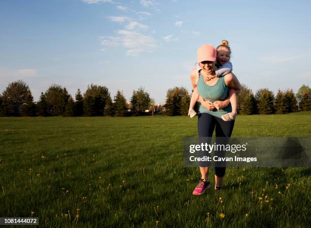 happy mother piggybacking daughter while exercising on grassy field against sky at park - madre-corriendo fotografías e imágenes de stock