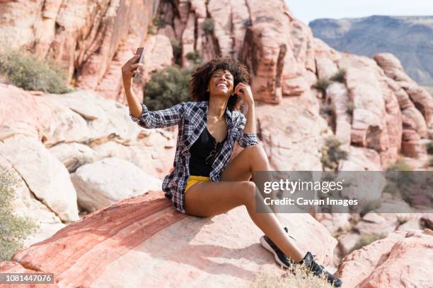 happy woman taking selfie while sitting on rock formation during sunny day - nevada hiking stock pictures, royalty-free photos & images