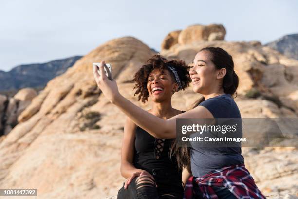 female friends taking selfie while sitting by rock formation against clear sky - nevada day stock pictures, royalty-free photos & images