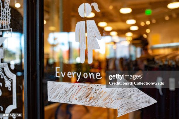 Close-up of hand drawn sign on a window depicting a stick figure which is half female and half male, with an arrow pointing towards an all gender...