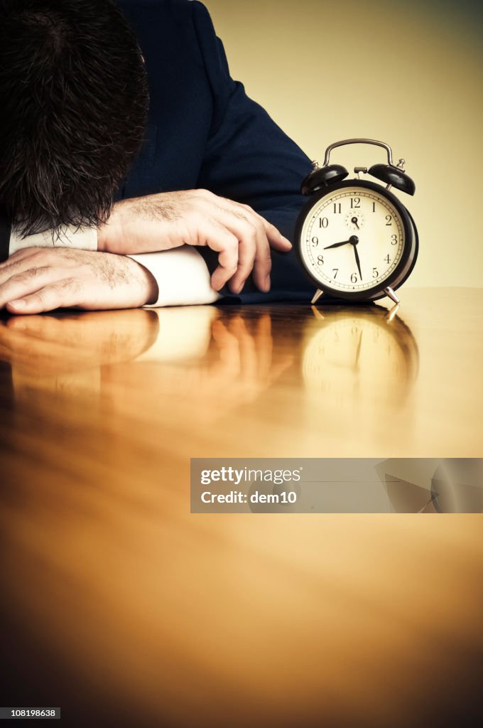 Businessman Resting Head on Desk Beside Alarm Clock