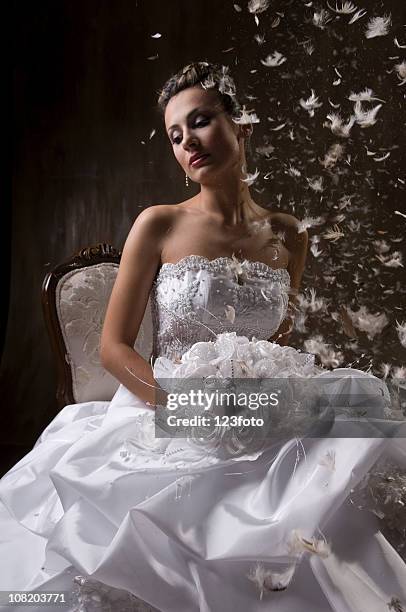 thoughtful bride sitting on chair with feathers floating around her - wedding dress stock pictures, royalty-free photos & images