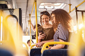 Two girls watching phone and smiling while standing on a bus.