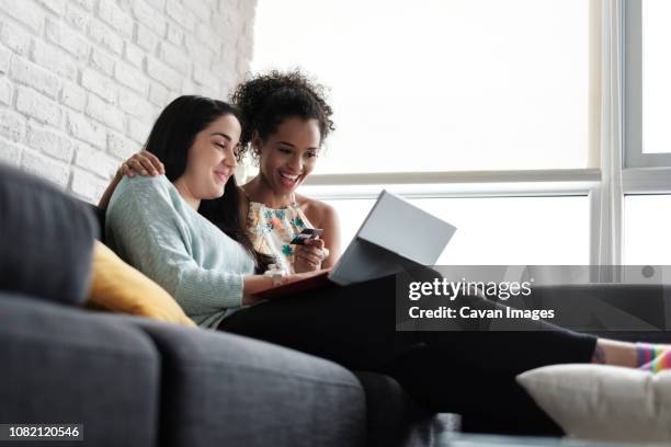 low angle view of happy lesbian couple doing online shopping while sitting on sofa at home - mobile banking stock-fotos und bilder