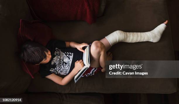 high angle view of boy with broken leg reading book while lying on sofa at home - gipsbein stock-fotos und bilder