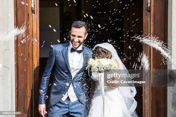 confetti throwing on happy newlywed couple standing at church entrance - front on groom and bride stock pictures, royalty-free photos & images