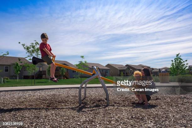 siblings playing on seesaw against sky at park during sunny day - see saw fotografías e imágenes de stock