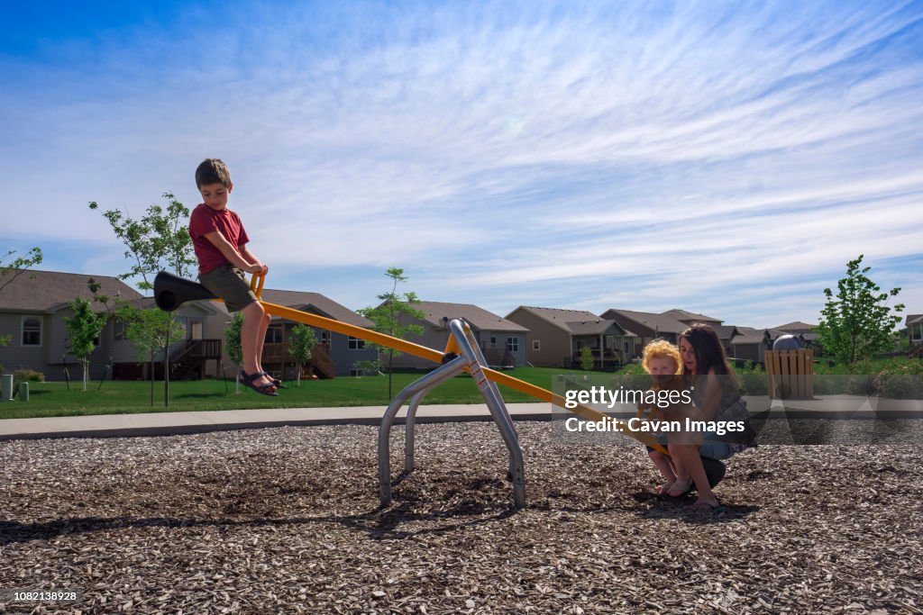 Siblings playing on seesaw against sky at park during sunny day