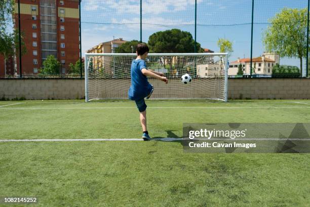 rear view of boy kicking soccer ball towards net on field - soccer goal stock-fotos und bilder