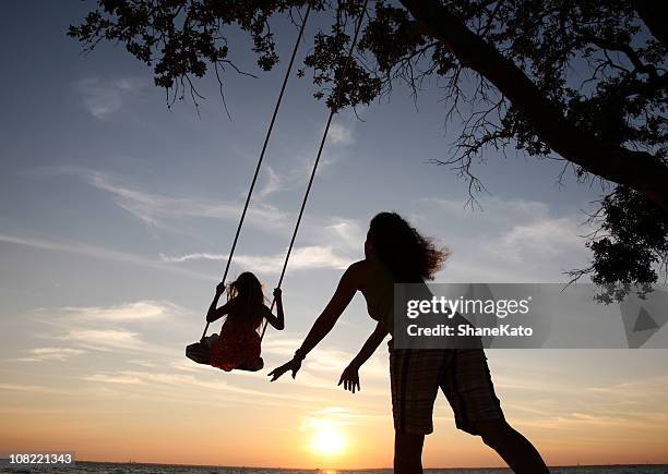 mom pushes daughter on swing set playing at sunset silhouette - oak tree silhouette stock pictures, royalty-free photos & images