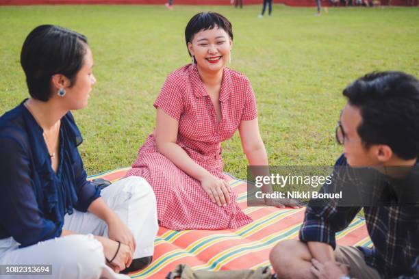 young woman smiling at two friends while sitting on a mat laid out on the grass - philippines friends stock pictures, royalty-free photos & images