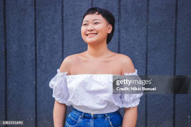 young woman standing in front of blue wall smiling and looking away - toma mediana fotografías e imágenes de stock