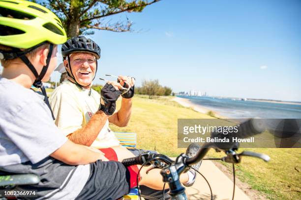 active australian senior takes his grandson out on their mountain bikes for a beach front ride - cycling helmet stock pictures, royalty-free photos & images