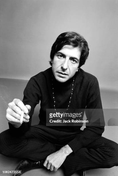 Portrait of Canadian poet, novelist, and musician Leonard Cohen , dressed in black, as he sits crossed legged on the floor and holds a cigarette in...